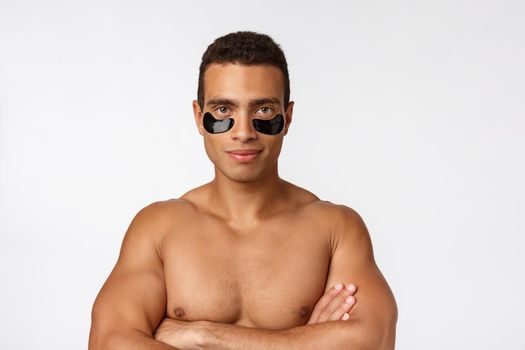 Attractive african young man with under eye black patches caring for his facial skin looking straight at camera on grey background