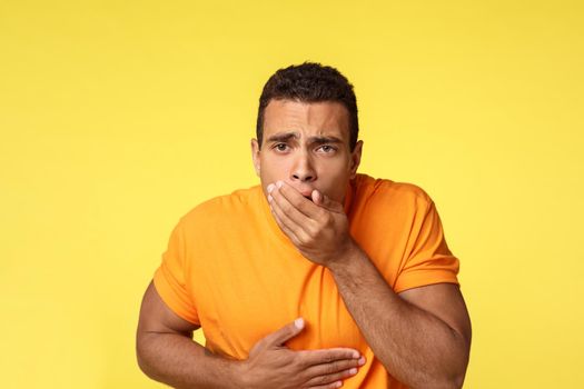 Masculine man want vomit, feeling sick and unwell, ate too much junk unhealthy food, touching stomach and cover mouth with palm as standing yellow background sick, puke from disgust