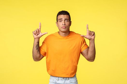 Sad gloomy young caucasian man in orange t-shirt frowning upset, pointing up offended or distressed, losing competition, feeling sadness and depression, standing frustrated over yellow background