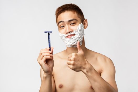 Beauty, people and hygiene concept. Pleased good-looking asian man recommend razor or shaving cream, show thumb-up and smiling pleased while face in cream, standing white background