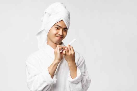 Beauty, spa and leisure concept. Portrait of silly good-looking asian man in bathrobe and bath towel, smiling pleased, polishing nails and make manicure, standing white background