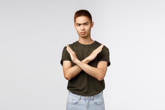 Serious-looking angry asian man make cross sign and frowning disappointed, saying no, disapprove and disagree, give negative feedback, prohibit and forbid, grey background