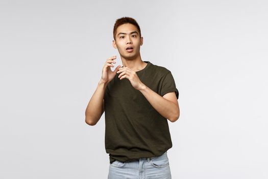 Portrait of startled young asian guy, male student gasping and jumping scared as someone appeared unexpected, raising hands up defense, stare terrified at camera, standing grey background