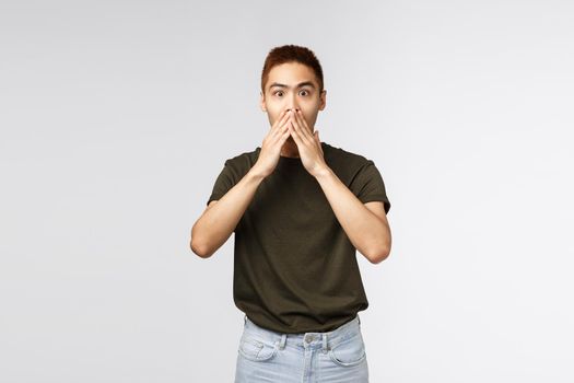 Surprised and amused asian man gasping, cover mouth and look camera impressed, express astonishment and happiness for friend, hear great news, standing grey background pleased