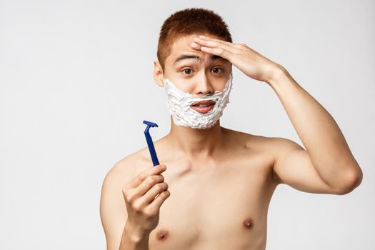 Beauty, people and hygiene concept. Confused cute asian teenage guy trying shave first time, feel little nervous, touching forehead, showing razor and apply shaving cream, white background