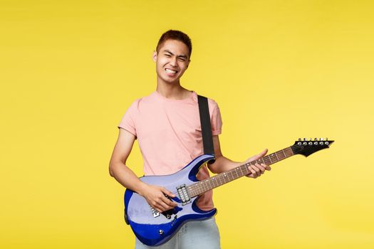 Lifestyle, leisure and youth concept. Lets jam. Carefree smiling asian guy playing in band, holding blue electric guitar, feel rock-n-roll start on stage, standing upbeat yellow background