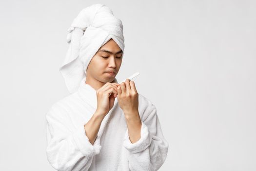 Beauty, spa and leisure concept. Cute asian man in bathrobe and bath tube taking care of personal hygiene, polishing nails, make own manicure during quarantine lockdown, white background