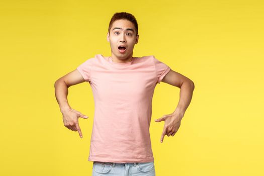 Portrait of overwhelmed, impressed asian hipster guy, taiwanese man showing advertisement, pointing fingers down, open mouth fascinated, showing friend promo, yellow background
