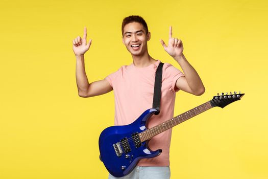 Lifestyle, leisure and youth concept. Happy enthusiastic handsome young asian male pointing fingers up, performing on stage, holding electric guitar and smiling, yellow background