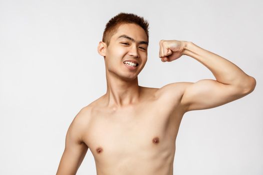 Workout, lifestyle and health concept. Strong proud handsome asian man, flex biceps and smiling pleased, become strong after exercises, attend gym, standing white background