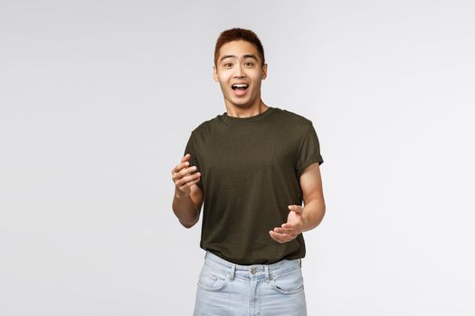 Portrait of surprised happy asian man, laughing and gasping amused, react to unexpected awesome news, express rejoice and happiness, standing grey background astonished
