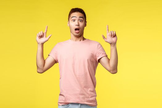 Lifestyle, people and summer vacation concept. Surprised, impressed asian male in pink t-shirt, pointing fingers up, open mouth astonished and amazed, showing new advertisement, cool product