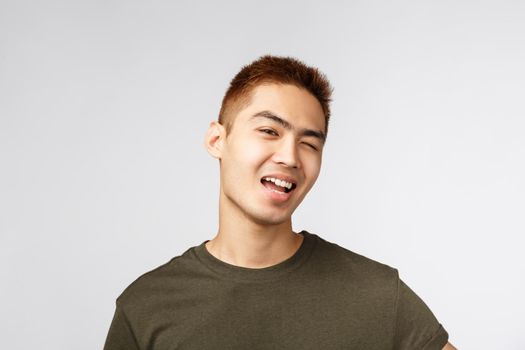 Portrait of good-looking sassy asian young male with proud ego, wink cheeky and coquettish, smiling please, standing grey background full of enthusiasm and positivity