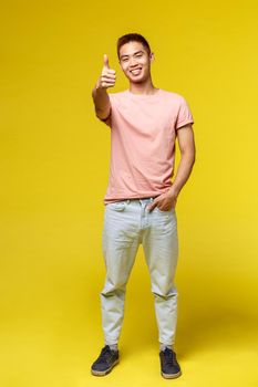 Lifestyle, education and people concept. Vertical portrait of stylish asian hipster guy, show thumb-up and smiling pleased, recommend product, approve and guarantee great quality, yellow background
