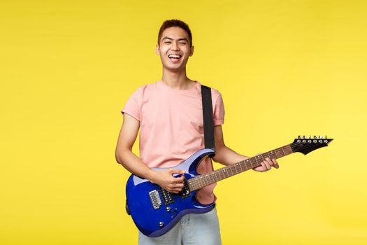 Lifestyle, leisure and youth concept. Happy asian male student playing in rock band, holding electric guitar and singing carefree with pleased smile, like perform on stage, yellow background