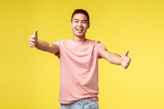 Lifestyle, travel and people concept. Portrait of cheerful, friendly asian male inviting for hug, reaching hands for embraces, stretch arms and smiling broadly, cuddling, standing yellow background