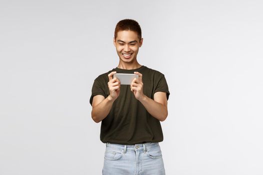 Technology, online lifestyle and communication concept. Happy pleased young asian male using mobile phone, smiling cheerful as playing new smartphone app, grin satisfied, grey background