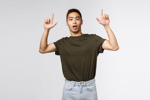 Portrait of shocked and nervous asian man look at camera surprised, pointing fingers up, showing something strange and concerning, asking question about item, grey background