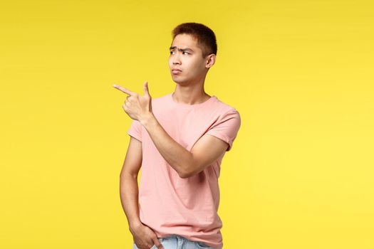 People, different emotions and lifestyle concept. Skeptical displeased asian guy in pink t-shirt, pointing finger left and look at something disappointing, dont like it, express judgement
