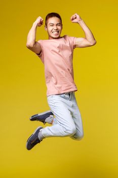 Summer vacation, education and lifestyle concept. Vertical shot of happy athletic asian guy, flex biceps and smiling upbeat, jumping from joy, triumphing, celebrating victory, yellow background