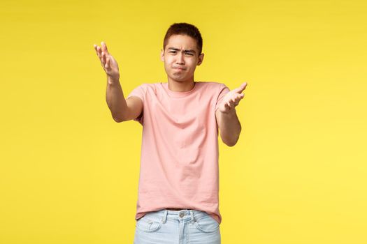Portrait of frustrated and disappointed asian guy indicating at something distressing and bad, grimacing complaining bad work, awful quality, judging, standing skeptical yellow background