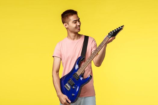 Lifestyle, leisure and youth concept. Cheerful handsome asian male student playing in band, tune electric guitar and smiling pleased, ready to jam on stage, perform new song, yellow background
