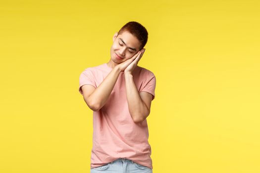 People, different emotions and lifestyle concept. Dreamy asian sleepy cute guy in pink t-shirt, dreaming about going summer vacation, lean on palms like falling asleep, smiling, yellow background