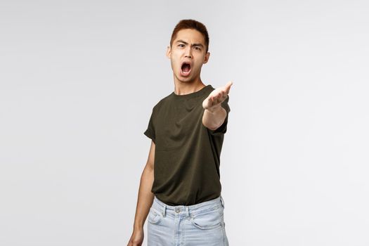 Portrait of angry complaining asian man pointing hand at camera with aggressive distressed look, disappointed arguing, received wrong parcel, being outraged standing grey background