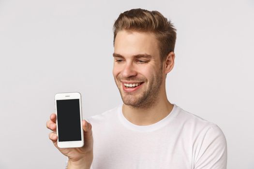 Joyful cute handsome blond bearded boyfriend in white t-shirt, holding smartphone, showing mobile display, smiling pleased, bragging with personal social media page, application score