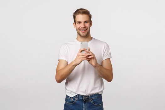 Excited, happy and cheerful blond stylish handsome man in white t-shirt, record concert on phone, taking picture, photoraphing, making good shot with smartphone camera, smiling thrilled