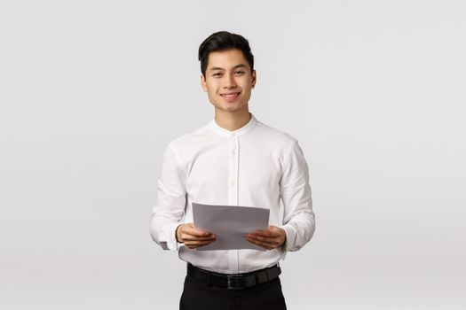 Handsome elegant asian young male coworker, businessman in white collar shirt, black pants, holding papers and smiling camera, successful entrepreneur prepare report, reading speech