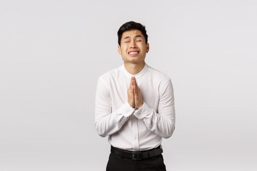 Worried, hopeful young asian male entrepreneur, businessman praying, begging boss give him second chance, close eyes clench teeth nervously supplicating, standing white background