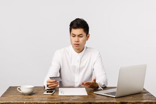 Distressed questioned and frustrated young asian guy sitting office at work, with documents and laptop, looking at credit card and complaining strange transactions, standing white background
