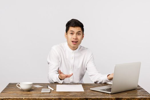 Frustrated and upset, distressed young asian office manager male, sitting table and spread hands sideways dismay, complaining, having failure, bad results, feeling regret and disappointment