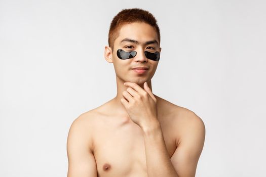 Beauty, people and lifestyle concept. Thoughtful enthusiastic, handsome naked asian man wearing eye-patches and smiling, have an idea, thinking, consider apply skincare mask, white background