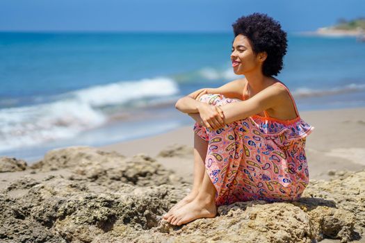 Happy black female sitting on the rocks on the beach looking at the sea smiling