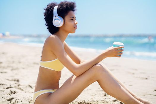 Black woman sitting on the beach with headphones, sipping a drink from a paper take-away cup.