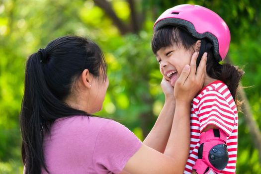 Asian mother helps daughter put on protective pads and safety helmet before practicing roller skating in the park. Exciting outdoor activities for kids.