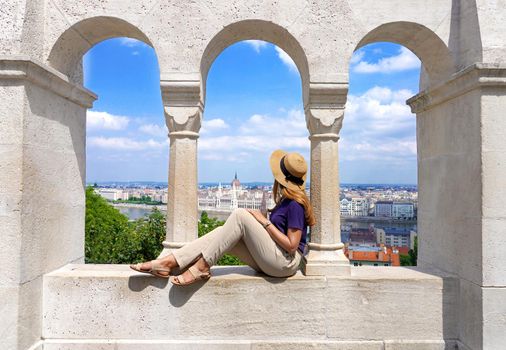 Beautiful young woman with hat sitting on wall looking at stunning cityscape of Budapest, Hungary