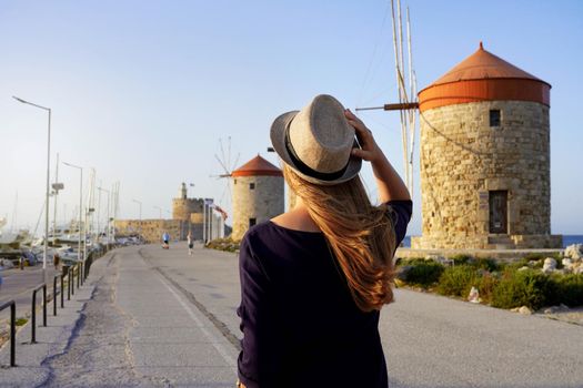 Visiting Rhodes in Greece. Back view of young traveler woman walking along promenade with old windmills of Rhodes, Europe. Sunset.