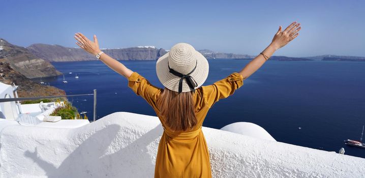 Europe travel vacation woman with arms up looking Santorini Caldera in Greece. Carefree tourist girl in European destination wearing fashion dress and hat.
