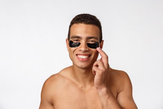 Attractive african young man with under eye black patches caring for his facial skin looking straight at camera on grey background
