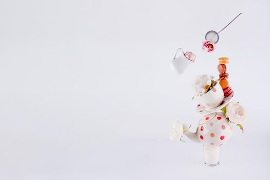 balancing structure of polka dot teapot, cup and saucers with macaroons and levitating milk jug and tea strainer blooming with flowers on white background