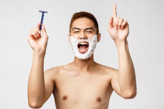 Beauty, people and hygiene concept. Portrait of enthusiastic asian naked man in bathroom, singing during shaving, holding razor and dancing as trying rich highest note, apply cream on face
