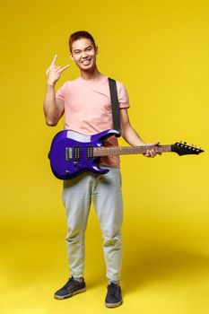 Lifestyle, leisure and youth concept. Vertical shot of enthusiastic teenage guy having fun playing electric guitar, show rock-n-roll sign and smiling, stand yellow background, play in band