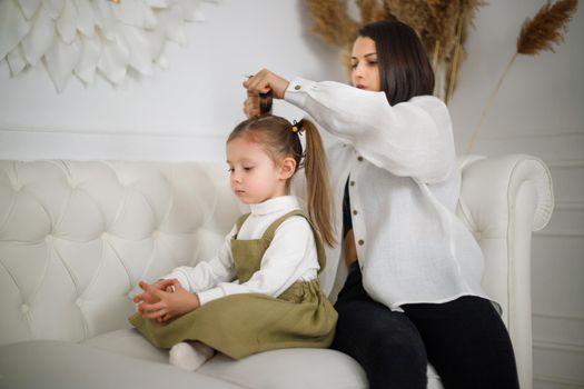 Focused mum collects daughters hair while sitting on the couch in luxury white interior