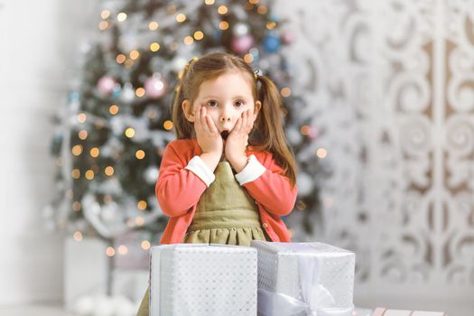 Emotional girl surprised by the abundance of New Year gifts by a Christmas tree in cozy room