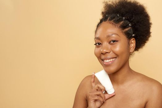 Lovely african american woman holding tube with moisturizer or mask mockup on beige background with copy space. Skin care, anti aging