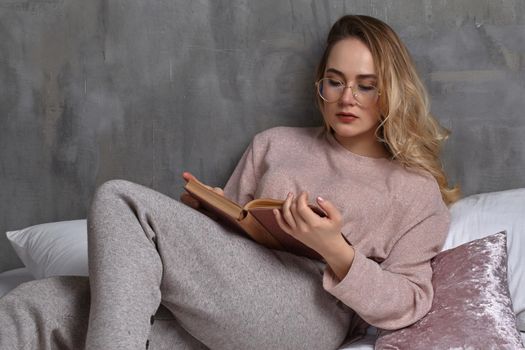 Blonde female in glasses, casual clothing is reclining on bed, reading book in bedroom. Student, blogger. Interior with gray wall. Close-up