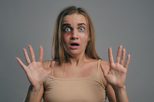 Blonde girl with bruises on her face, trying to protect herself, posing on gray studio background. Domestic violence, abuse. Close-up, copy space.
